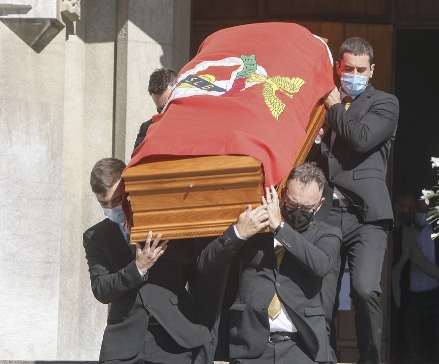 Quatro homens vestidos de preto e máscara carregam caixão de madeira coberto com bandeira SLB